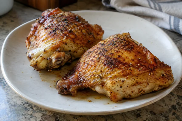 Two browned chicken thighs with skin and bone on an off white plate